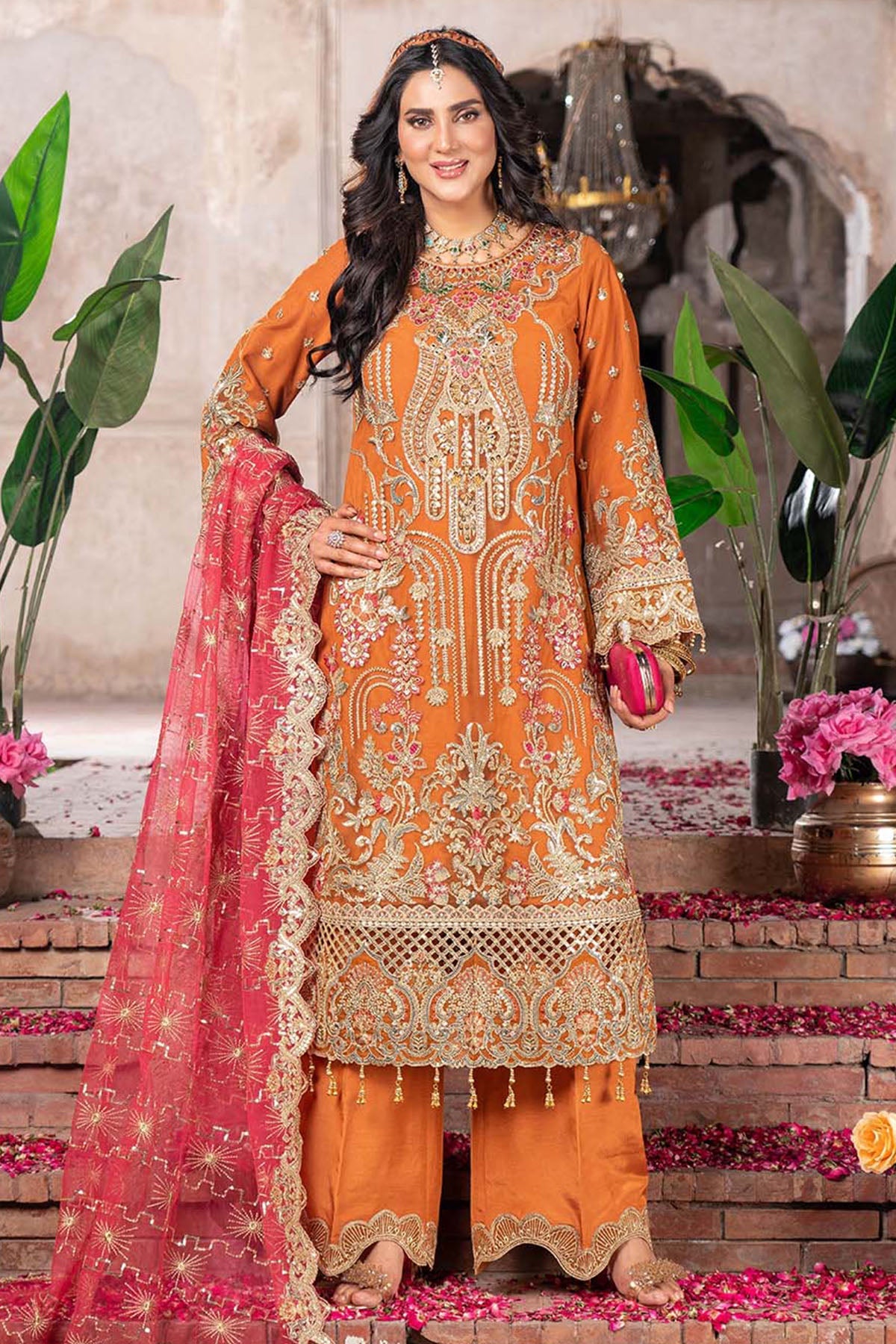 Woman in an orange embroidered outfit with a pink dupatta standing on a decorative step.