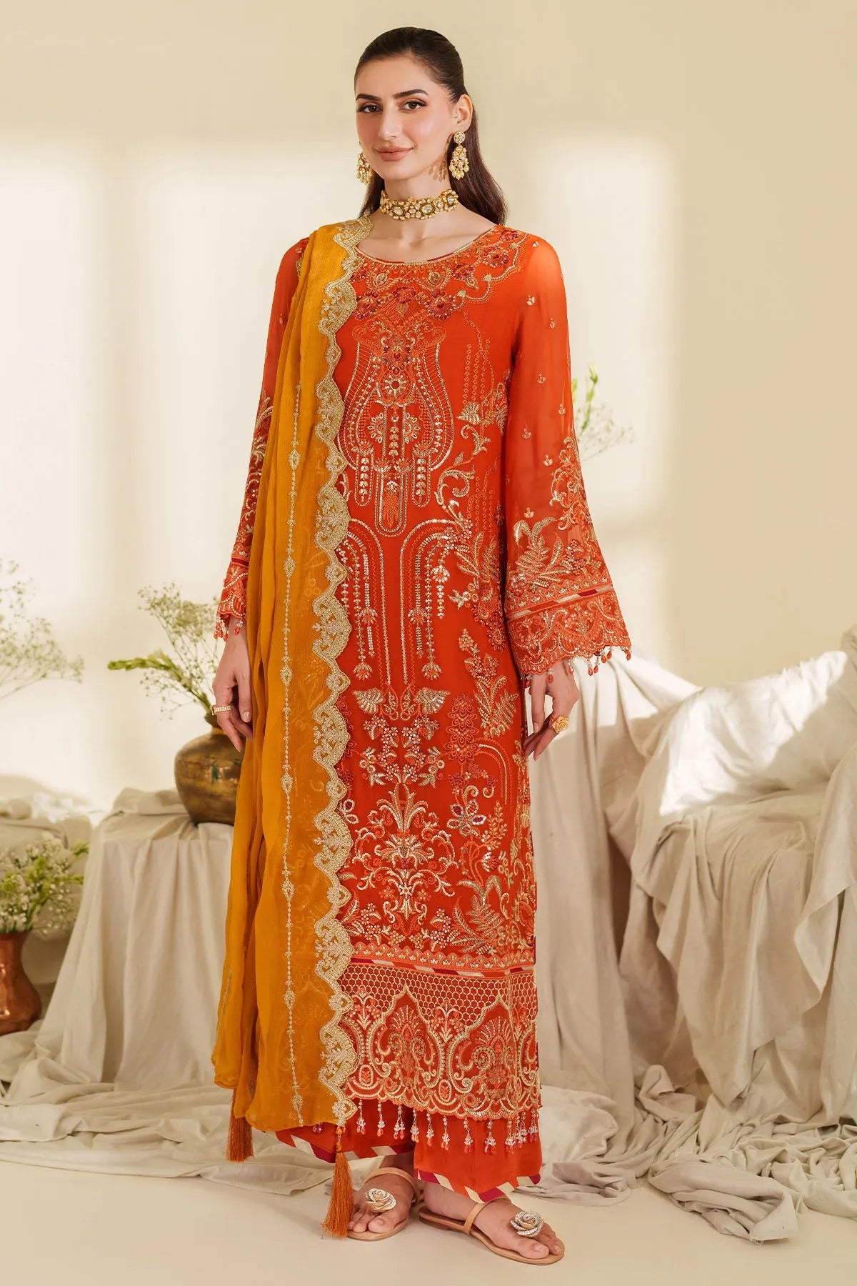 Woman wearing an orange embroidered outfit with a yellow dupatta in a softly lit room.