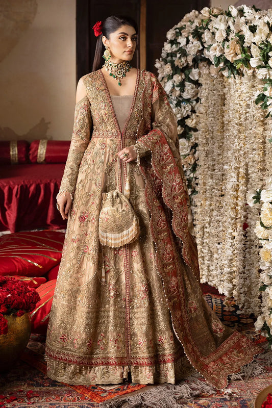 A woman wearing a traditional, embroidered beige gown with a floral pattern, paired with a dupatta and organza sleeves, standing in a decorative setting with floral arrangements in the background.