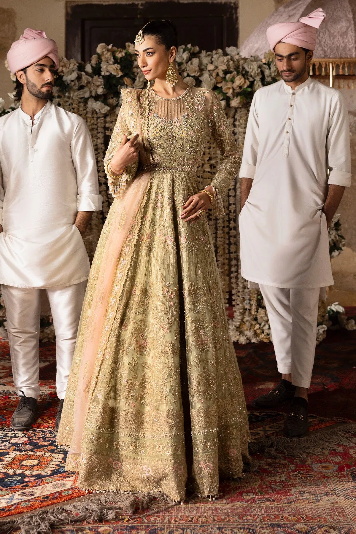 A woman in a beige and gold embroidered bridal gown with netted details and organza borders, accompanied by men in white traditional attire.